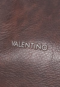 Rjava usnjena površina z teksturirano strukturo, ki vsebuje izrazit srebrni logotip "VALENTINO". Preprost dizajn poudarja bogato barvo.