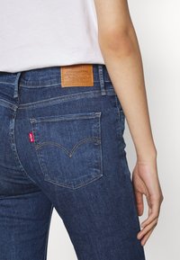Tmavě modré džíny z denimu s texturovaným povrchem. Obsahují hnědou koženou nášivku na zadním pásu a červené logo Levi's na zadní kapse.