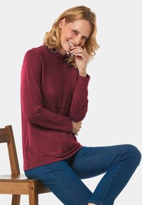 Burgunderfarbenes Langarmoberteil mit hohem Kragen, das eine weiche Textur und einen geraden Schnitt aufweist. Das Model sitzt auf einem Holzstuhl und trägt eine blaue Jeans.