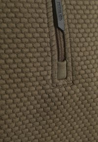 Olijfgroene textuurstof met een honingraatpatroon, voorzien van een bruine rits en een klein label met de tekst "PETROL IND."