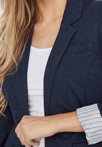 Blazer blu scuro con vestibilità su misura, con rever eleganti e un discreto dettaglio sulla tasca. Sotto, una maglietta bianca con una manica a righe in vista.