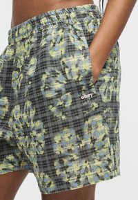 Άτομο που φοράει σορτς με ελαστική μέση σε πράσινο και μπλε καρό με ανθισμένο μοτίβο, χέρι στην πλαϊνή τσέπη, το λογότυπο «obey» κεντημένο κοντά στην τσέπη.