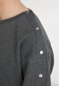 Grå sweatshirt laget av mykt stoff, med metalltrykknapper på skulderen og en avslappet passform. Teksturert overflate synlig.