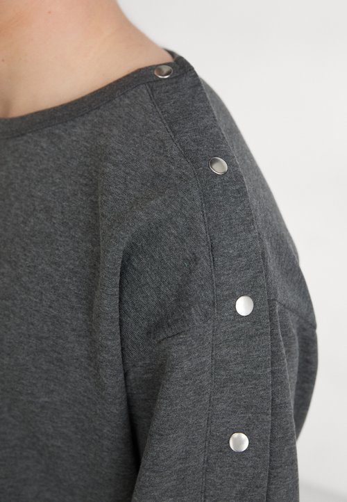 Pilkas megztinis, pagamintas iš minkštos medžiagos, su metaliniais užsegimais ant peties ir laisvu kirpimu. Matoma tekstūruota paviršius.