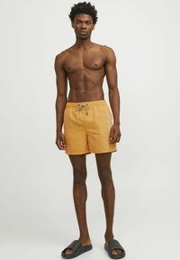 Bañador naranja con cintura elástica y cordón ajustable. Presenta bolsillos laterales y un logo impreso en blanco en el lado izquierdo. Chanclas negras lisas.