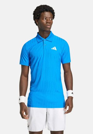 Mann trägt ein leuchtend blaues Poloshirt, weiße Shorts und weiße Armbänder, steht und schaut seitlich vor einem schlichten Hintergrund.