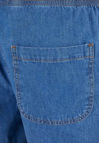 Džinsiniai šortai su elastine juosta, turintys vieną užpakalinį kišenę, mėlynos spalvos ir ryškiai oranžinė siuvinėjimo linija aplink kišenės kraštus.