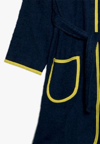 Ναυτικό μπλε μπουρνούζι φτιαγμένο από υφασμάτινη υφή, που διαθέτει ζώνη, δύο τσέπες με κίτρινες λεπτομέρειες και αντίθετη κίτρινη κοπή στα μανίκια και τις άκρες.