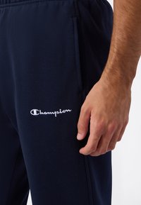 Primer plano de la mano de una persona descansando al lado de un pantalón de chándal azul marino con un logotipo de Champion bordado en blanco cerca del bolsillo.