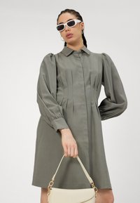 Rochie verde khaki cu guler, mâneci lungi bufante, față pliată și o siluetă ajustată. Asortată cu o geantă albă.