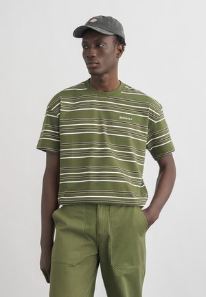 Hombre con una camisa de manga corta a rayas verdes y blancas, pantalones verde oliva y una gorra de béisbol gris oscuro, de pie con una mano en el bolsillo.