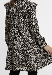 Robe à motifs noir et crème avec des détails volantés aux épaules, jupe à volants, manches longues et une silhouette fluide.