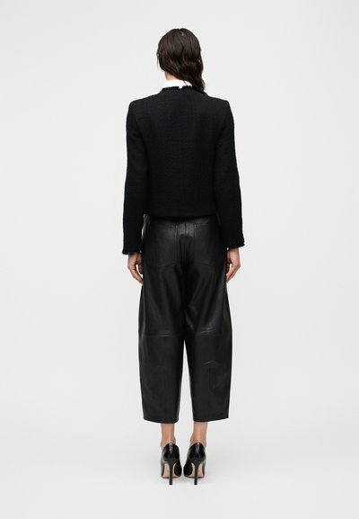 Μαύρο cropped μπουφάν από υφασμα με υφή, συνδυασμένο με υπερμεγέθη μαύρα δερμάτινα παντελόνια. Το μοντέλο φοράει μαύρα παπούτσια με ψηλό τακούνι.