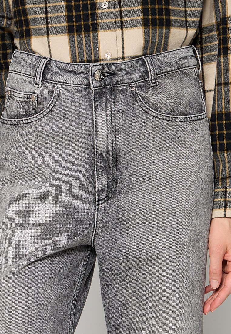 Szare dżinsy z wysokim stanem, pięć kieszeni, z widocznymi szwami; noszone z beżową i czarną koszulą w kratkę.