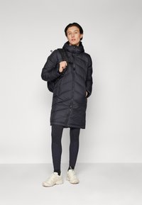 Svart pufferjacka med dragkedja fram, huva och diagonalt kviltat mönster, kombinerad med mörka leggings och ljusa sneakers, med en ryggsäck.