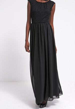 Femme portant une robe noire sans manches avec un corsage texturé et une jupe fluide en chiffon, associée à des escarpins noirs fermés sur fond blanc.