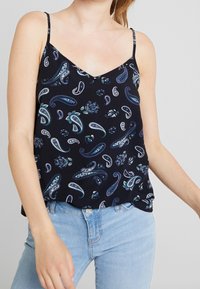 Femme portant un haut sans manches noir avec des motifs paisley bleus et blancs et un jean bleu clair, sur fond uni clair.