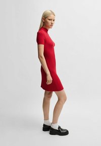 Rotes geripptes Kleid mit kurzen Ärmeln und hohem Halsauschnitt, körperbetont geschnitten und endet über dem Knie. Schwarze Plateauschuhe und weiße Socken.