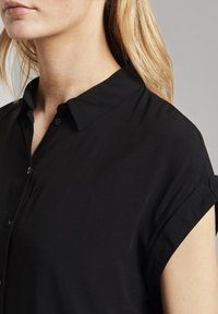 Primo piano di una donna bionda che indossa una camicia nera a maniche corte con colletto su uno sfondo grigio uniforme.