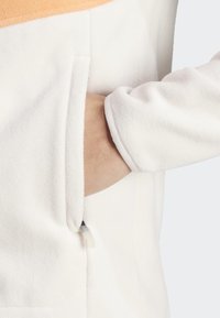 Chaqueta de forro polar blanca con una sección superior naranja, mostrando una mano alcanzando un bolsillo lateral. El material es suave con una textura lisa.