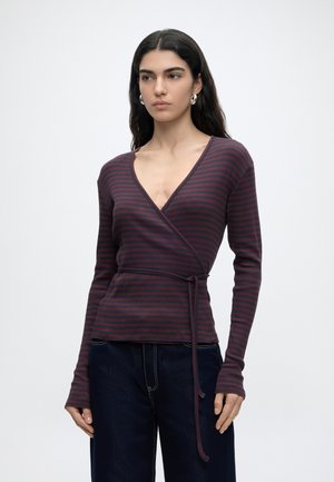 Femme aux longs cheveux foncés portant des boucles d'oreilles créoles argentées, un haut cache-cœur à manches longues rayé violet foncé et rouge avec ceinture nouée, et un pantalon foncé.