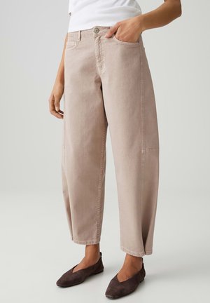 Frau trägt beige, hochgeschnittene, locker sitzende Hose mit vorderen Taschen und braunen flachen Schuhen, steht vor einem einfarbigen hellen Hintergrund.