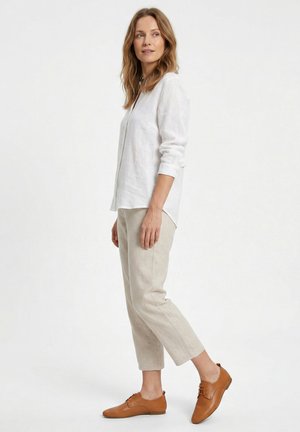 Vrouw staande in beige cropped broek, wit shirt met lange mouwen en lichtbruine veterschoenen, zijwaarts kijkend tegen een effen witte achtergrond.