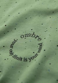 Grøn bomulds t-shirt med et cirkulært sort tekstdesign, der har sætningen "din eneste grænse er dit sind," samt prikker som accent.