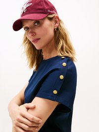 Maglietta blu navy con vestibilità rilassata, caratterizzata da bottoni dorati sulla spalla. Orecchini a cerchio dorati e cappellino da baseball bordeaux con testo bianco.