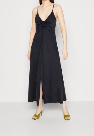 Kobieta w czarnej, bezrękawnikowej sukience maxi z rozcięciem z przodu i marszczeniem, w zestawieniu z oliwkowozielonymi, odkrytymi sandałami na obcasie.