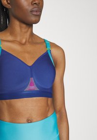 Sostén deportivo inalámbrico en azul profundo con copas acolchadas, que presenta tirantes ajustables en verde azulado y un detalle de acento rosa en el centro.