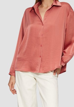 Vrouw die een losse, lange mouwen, poederroze blouse draagt met witte broek, staand tegen een effen lichtgrijze achtergrond.