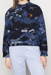 Marinblå sweatshirt med ett kamouflage mönster i ljusare blå och grå nyanser, ribbade muddar och en liten orange logotyp på bröstet.