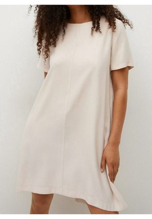 Vrouw in een losse, beige jurk met korte mouwen die voor een egale witte achtergrond staat en met één hand de zoom van de jurk vasthoudt.