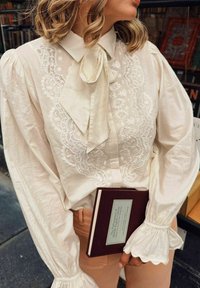 White blouse with intricate embroidery and a bow tie at the collar. Puff sleeves with ruffled cuffs. Holding a brown book with a green label.