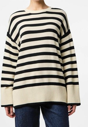 Vrouw die een loszittende beige trui met zwarte horizontale strepen draagt, gecombineerd met donkerblauwe spijkerbroek, staand tegen een effen achtergrond.