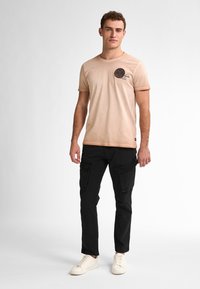 Béžové bavlněné tričko s kulatým potiskem, v kombinaci s černými cargo kalhotami s více kapsami. Model má na sobě bílé tenisky.