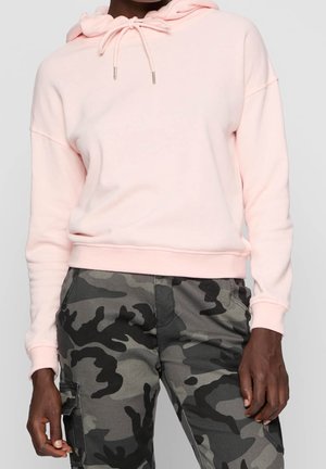 Persoon draagt een lichtroze hoodie met trekkoorden en grijs-zwarte camouflage cargobroek, staand met armen ontspannen langs het lichaam.