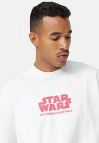 Valkoinen puuvillapaita, jossa on näyttävää punaista tekstiä: "TÄHTIEN SOTA: IMPERIUMI ISKEE TAKAS." Lyhyet hihat ja pyöreä kaula-aukko.