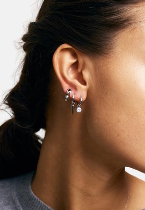 Sievietes auss ar trim sudraba lokveida auskariem: viens ar akmentiņiem, viens ar kodolīgu dekoru, un viens ar caurspīdīgu dārgakmens rotājumu.