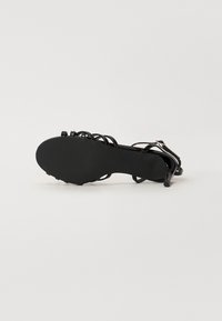 Sandale à talon noire à lanières, couchée sur le côté, montrant la semelle lisse et le talon fin de hauteur moyenne sur un fond blanc.