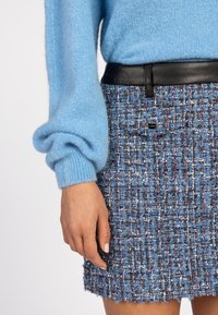 Handen rusten naast het lichaam, gekleed in een lichtblauwe fuzzy trui en een gestructureerde blauw en multicolor tweed rok met een zwarte leren riem.