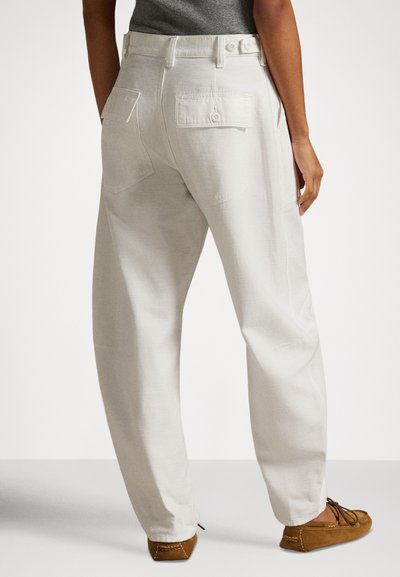 Person som har på seg løstsittende hvite bukser med lommepatcher og brune mokkasinsko, stående mot en ensfarget bakgrunn.