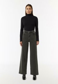 Czarna bluzka z golfem oraz zielone, wysokie spodnie o szerokich nogawkach, wykonane z denimu, z przednimi kieszeniami i zapięciem na guzik. Czarne obuwie.