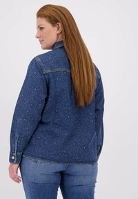 Kvinne med langt rødt hår iført en blå denimjakke prydet med små hvite nagler, kombinert med lyseblå jeans, vendt sidelengs.