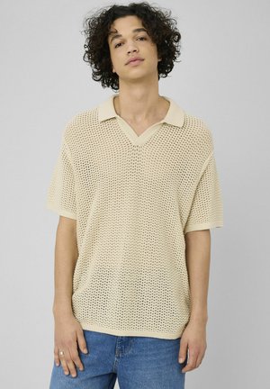 Jeune personne aux cheveux bouclés portant un polo beige en maille à manches courtes et un jean bleu, debout devant un fond clair uni.