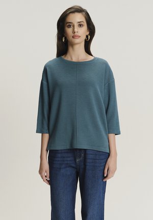 Turkusowy, prążkowany sweatshirt z okrągłym dekoltem, rękawami o długości trzech czwartych i luźnym krojem, połączony z ciemnoniebieskimi dżinsami.