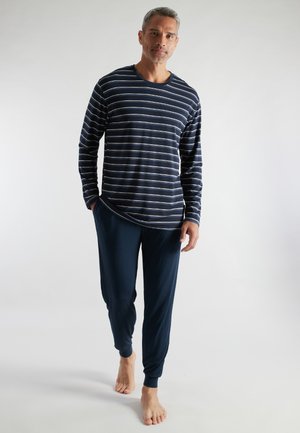 Mann läuft barfuß und trägt ein marineblau-gestreiftes Langarmshirt sowie passende marineblaue Hosen vor einem schlichten Hintergrund.