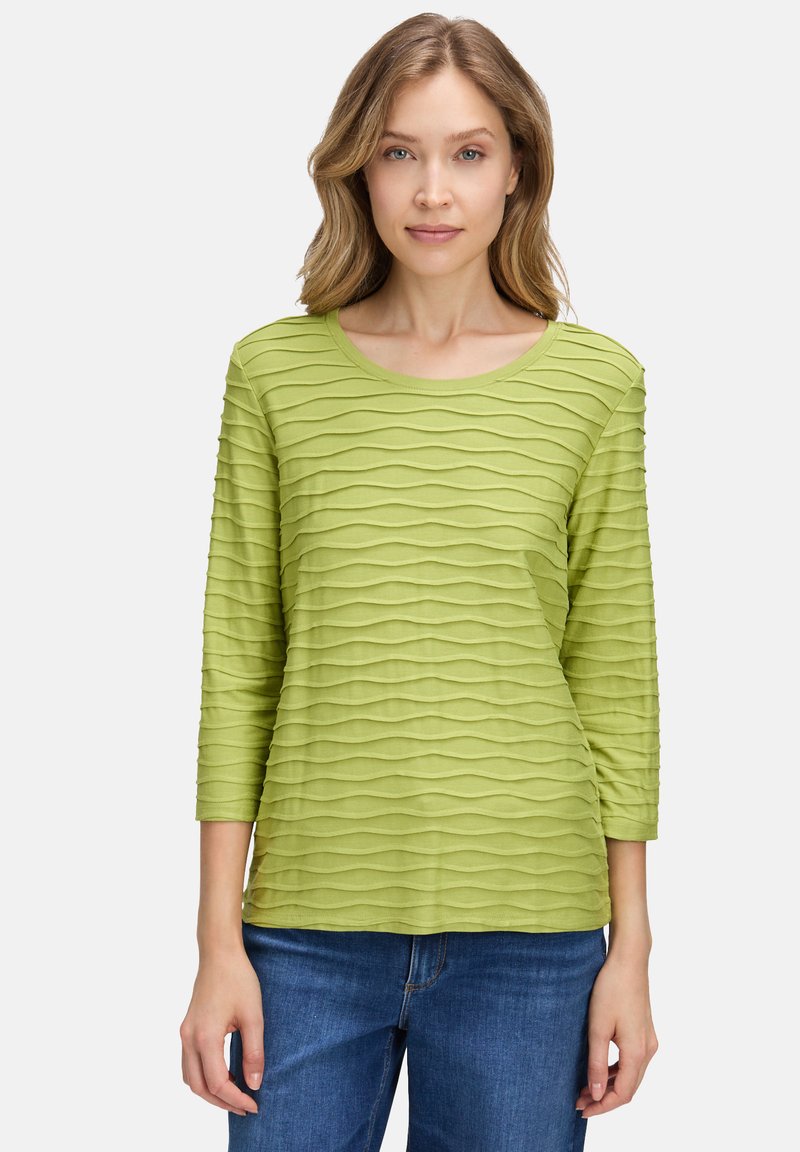 Grünes, strukturiertes Shirt mit langen Ärmeln und Wellenmustern, rundem Halsausschnitt und Dreiviertelärmeln, kombiniert mit blauen Jeans.