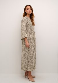 Long, patterned dress in beige with a wave-like design. Features a gathered waist and three-quarter sleeves. Paired with light-colored heels.